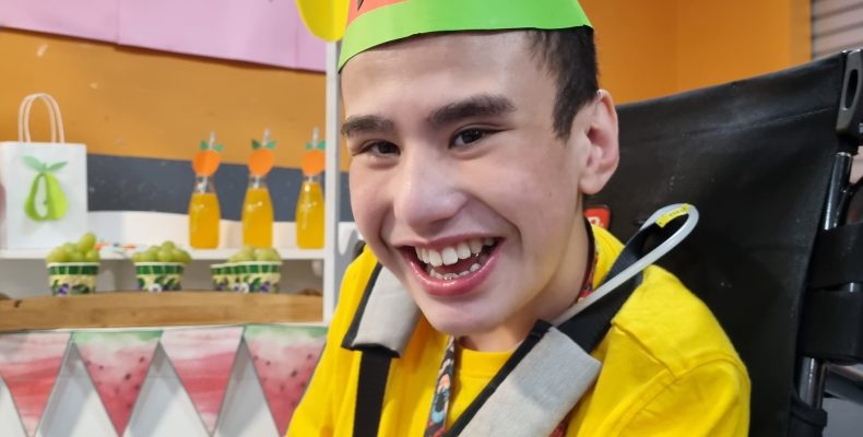 Image of a boy in a wheelchair, smiling with a fruit party hat on